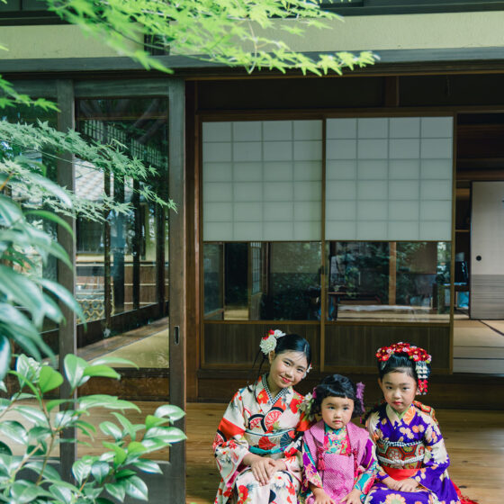 七五三用アンティーク着物姿の三姉妹が日本家屋の縁側で緑を背景に撮影するシーン